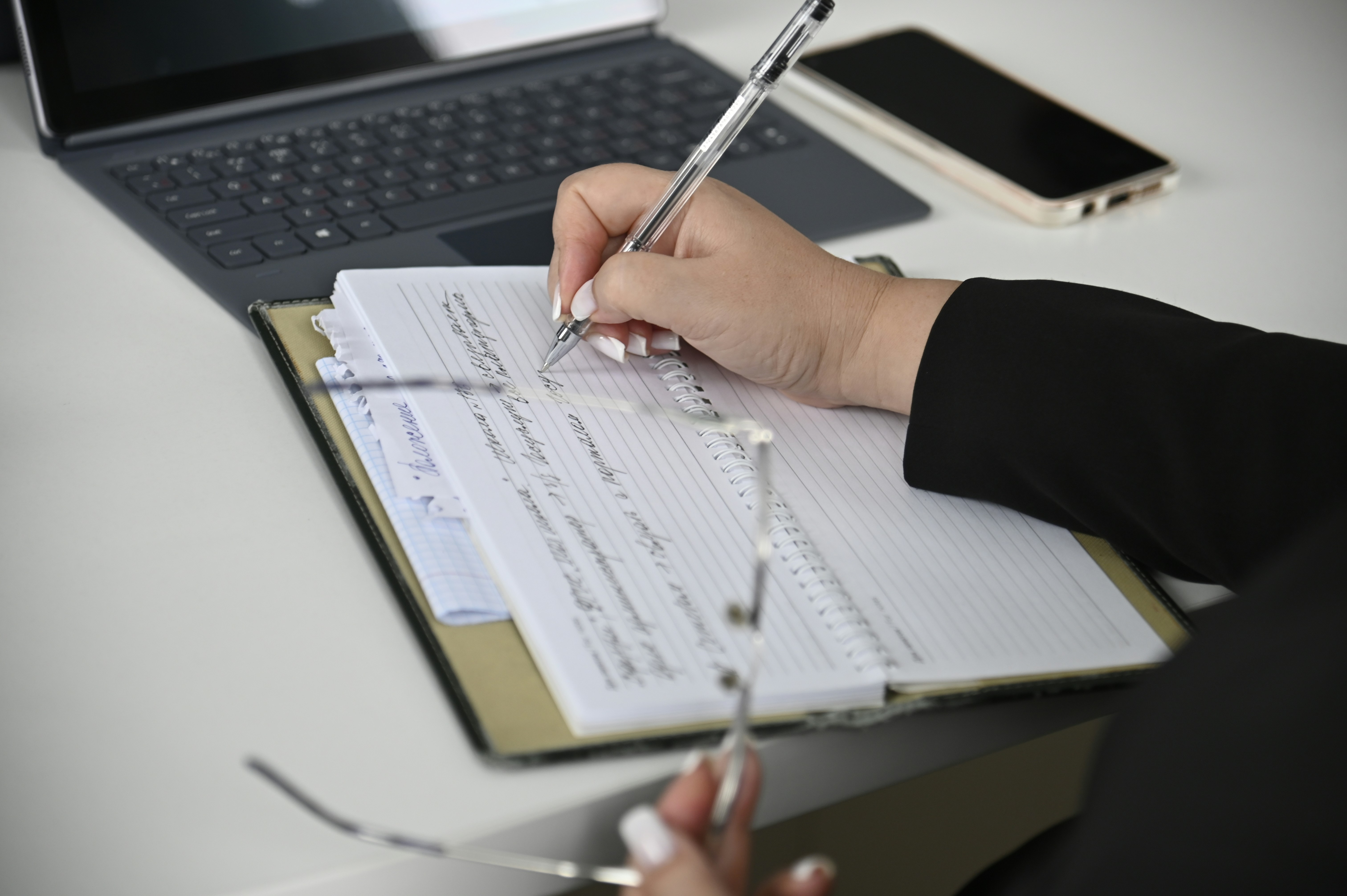 Close-up of hand writing notes with pen on notebook with laptop in background. Shows careful document review and analysis, expressing Working Draft's uncertainty and need for cautious approach